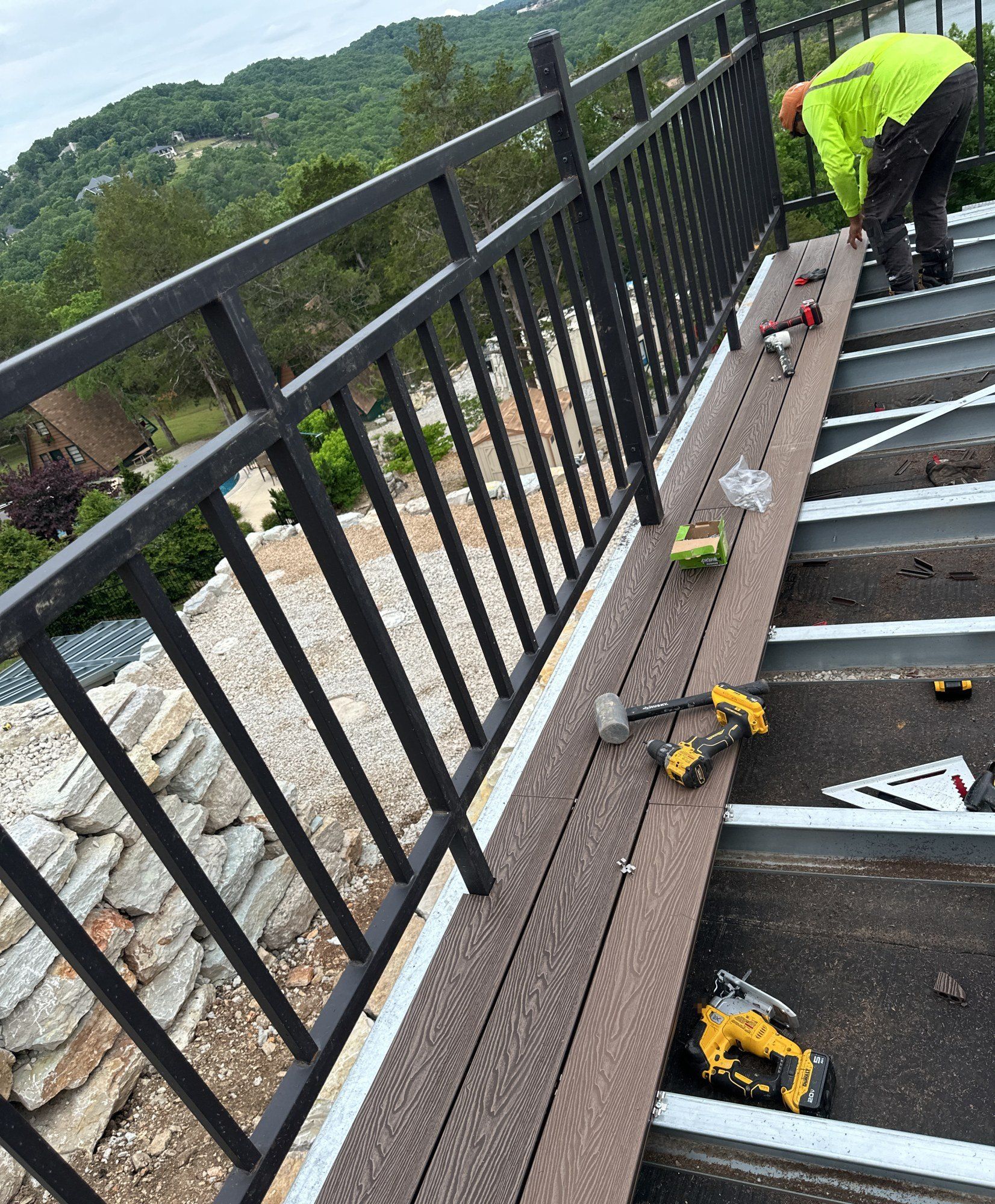 Worker laying wood-look deck boards across the steel deck frame beside a metal balustrade, hills behind, 26 May 2026
