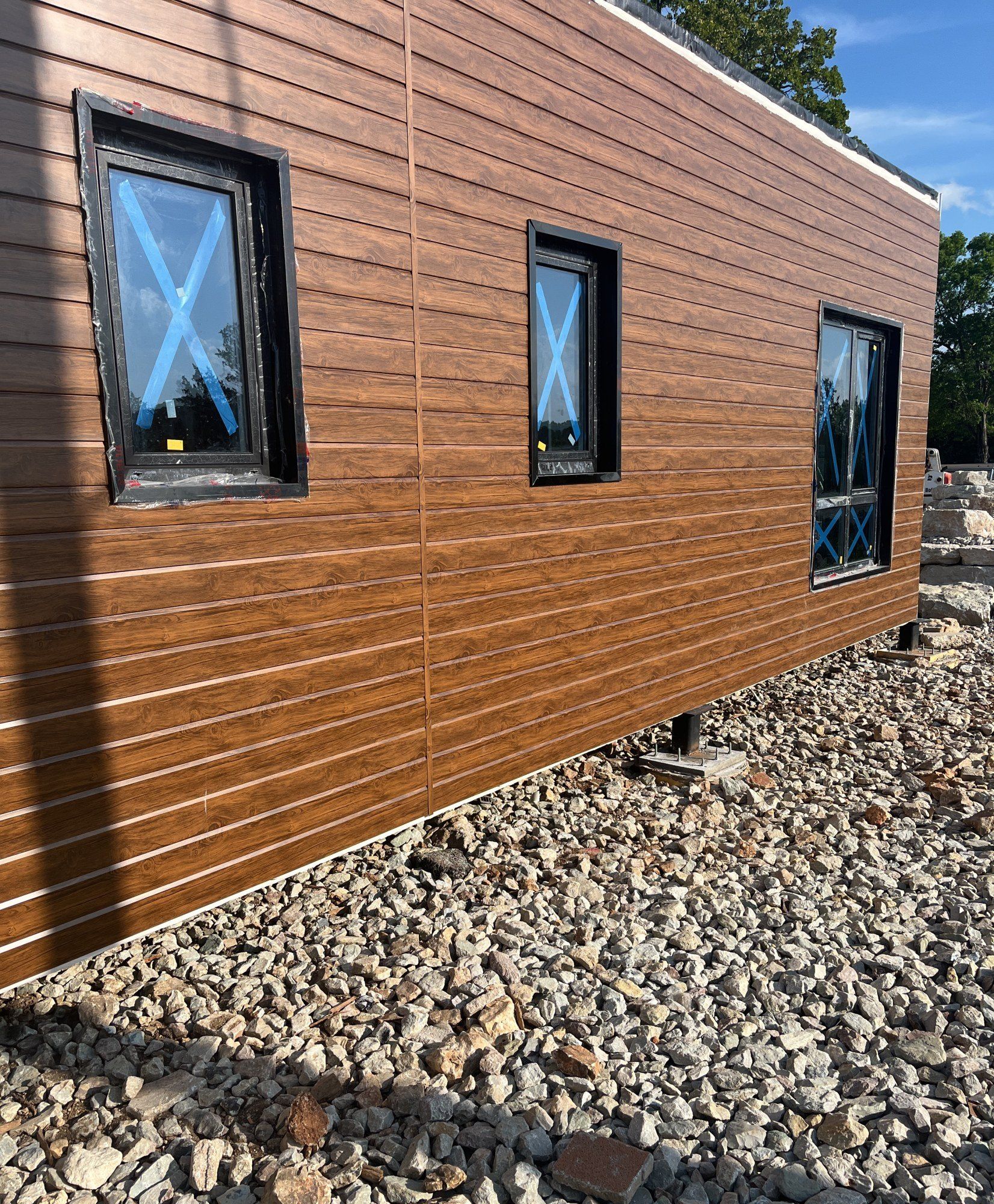 Exterior elevation in horizontal wood-look cladding with black-framed windows above a gravel bed, 18 May 2026