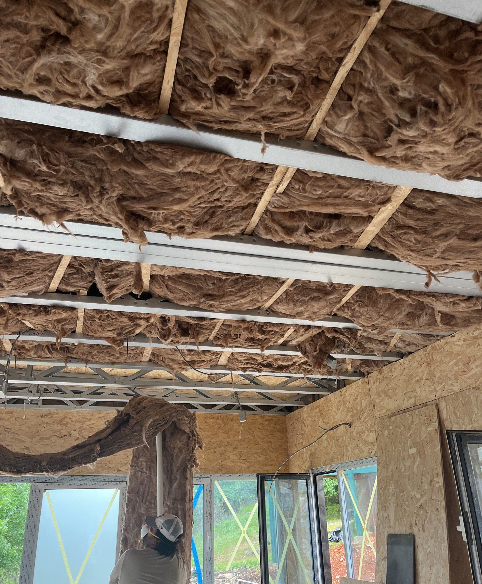 Insulation batts fitted between steel roof members above a sheathed ceiling, taped windows below, 27 May 2026