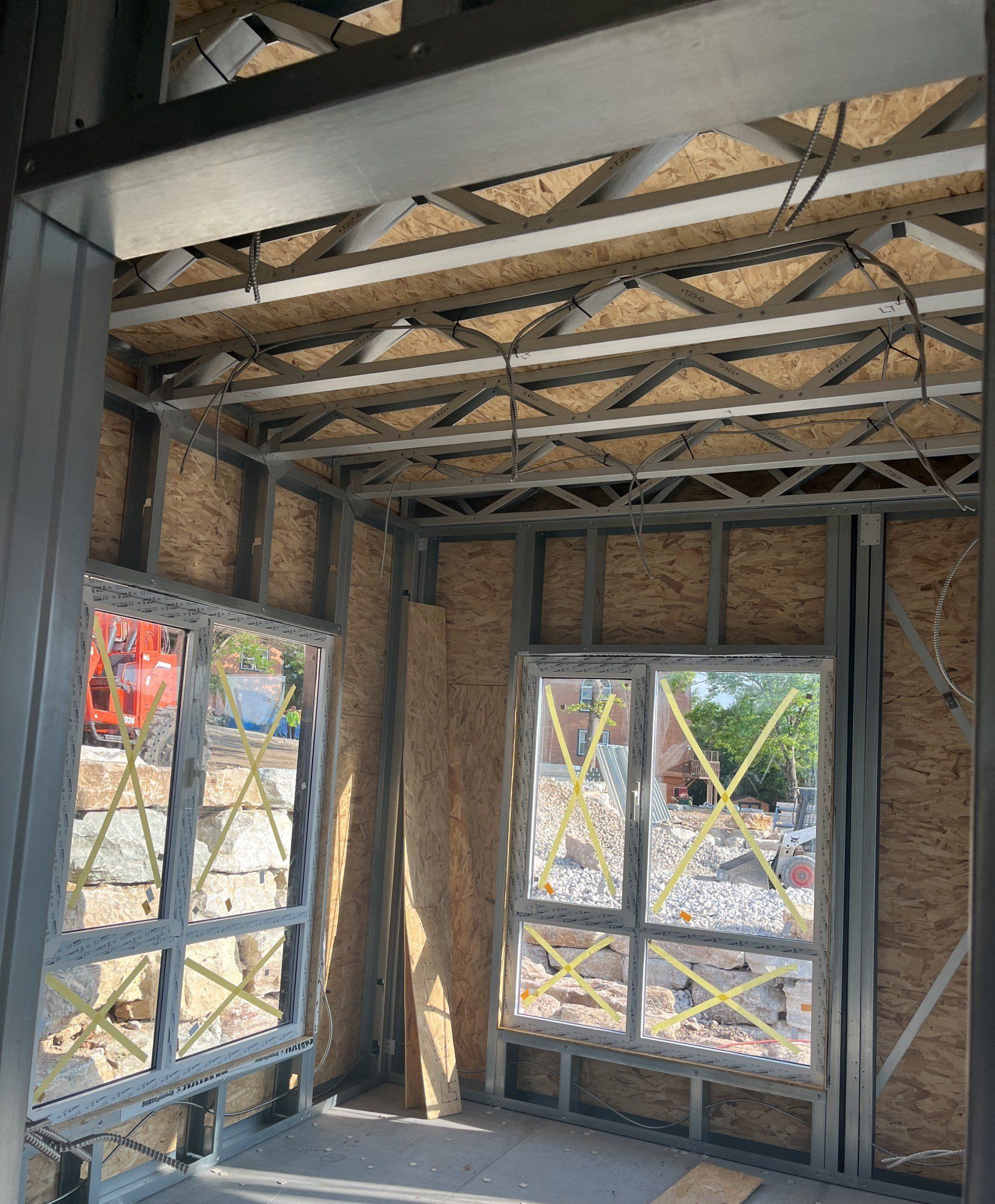 Corner of a room framed in steel with roof trusses overhead and taped window openings looking onto the site, 18 May 2026
