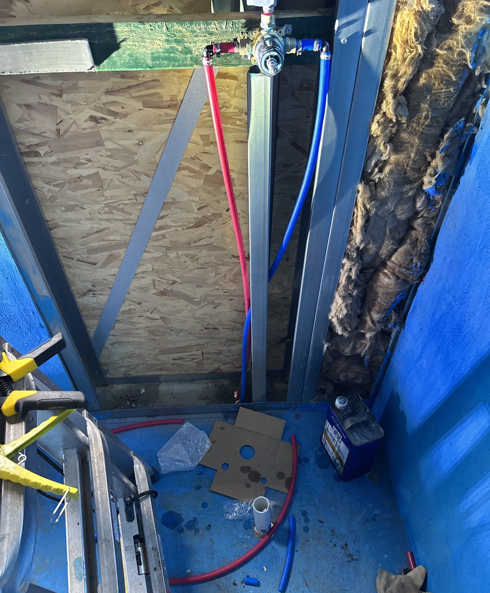 Plumbing manifold with red and blue pipe runs fixed against sheathing behind a blue wall membrane, 28 May 2026