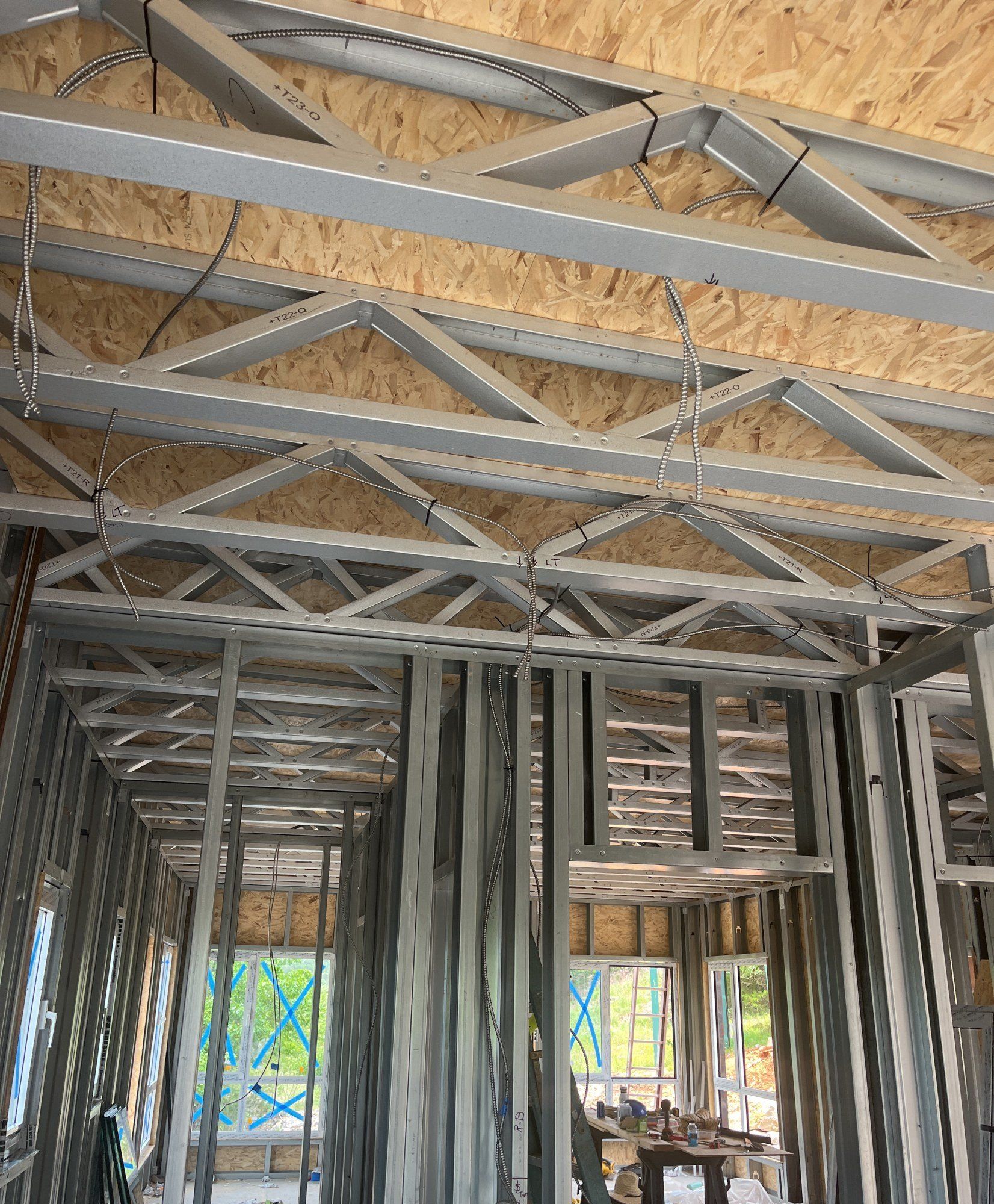 Interior view of steel roof trusses and steel wall studs with sheathing board between them, windows taped beyond, 18 May 2026