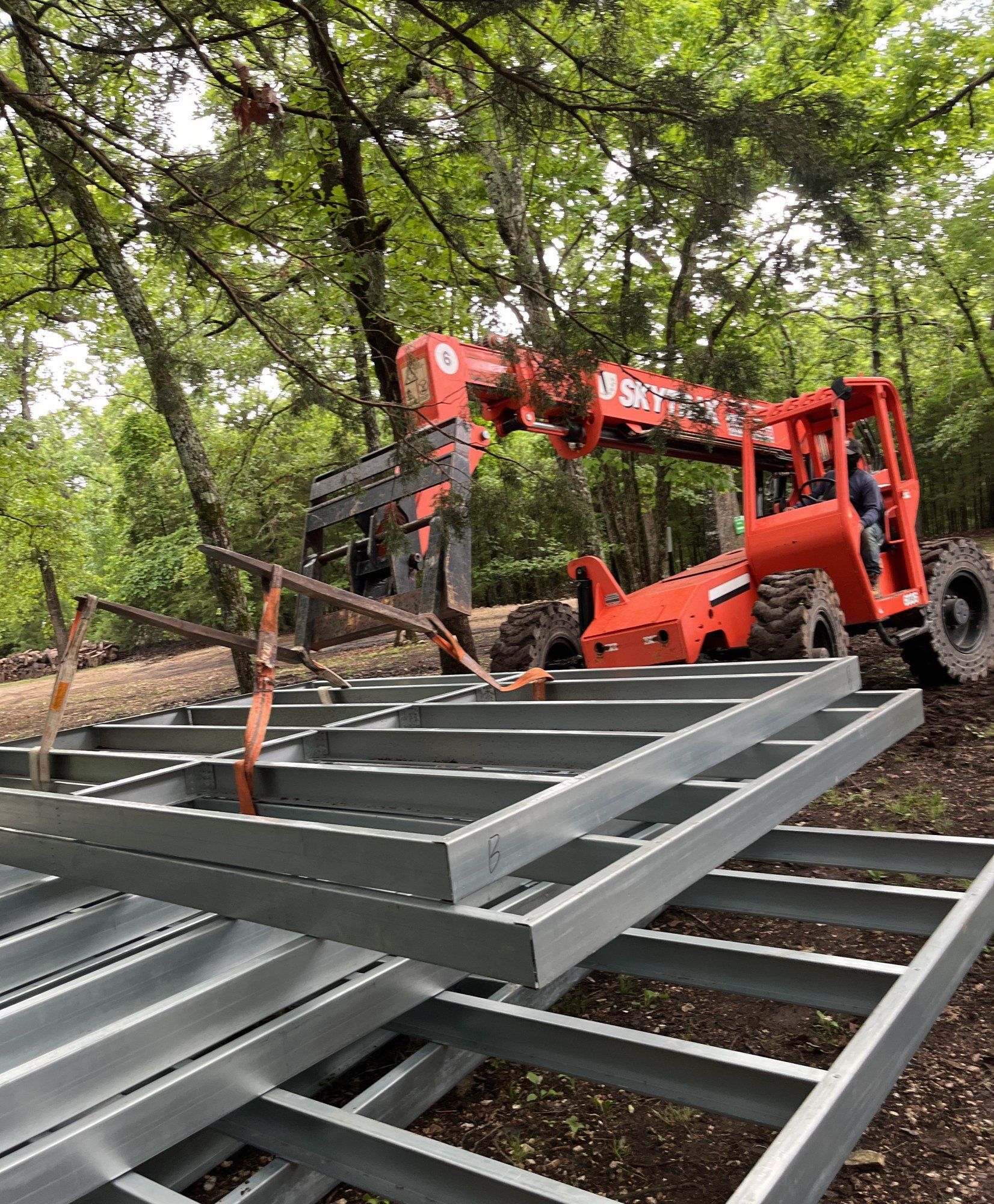 Telehandler lifting an assembled galvanized steel deck frame with lifting straps, wooded site behind, 20 May 2026