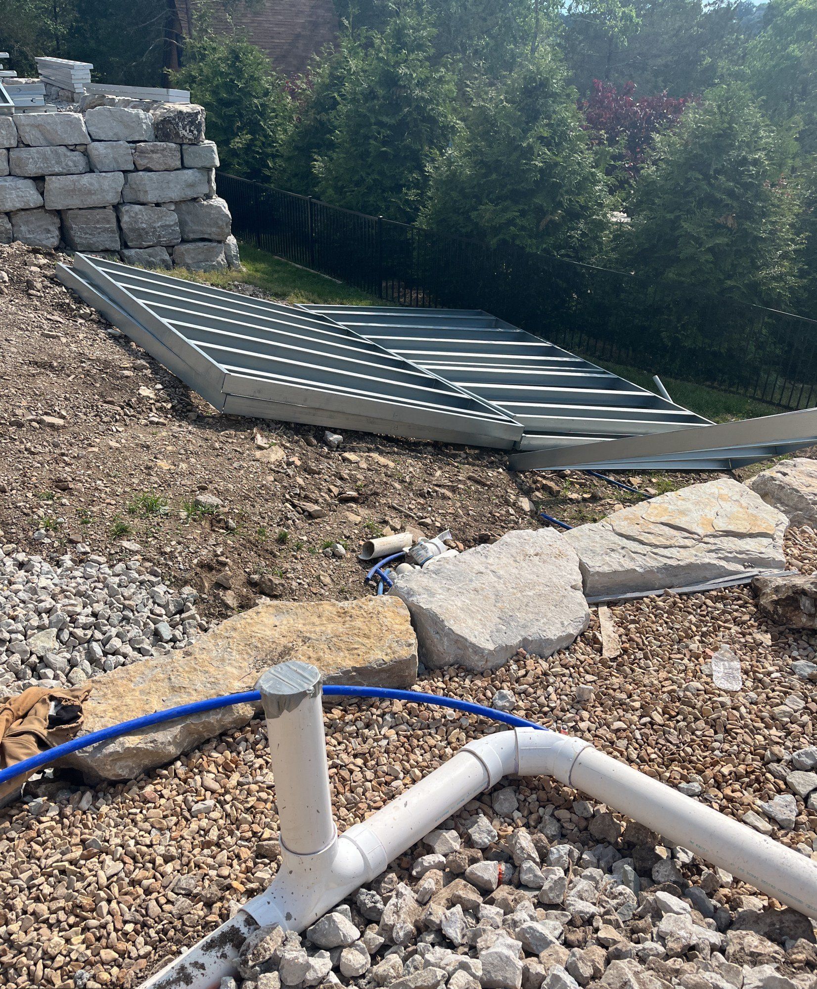 White drainage pipe and blue water pipe running through a gravel bed beside stacked steel deck frames and a stone retaining wall, 15 May 2026