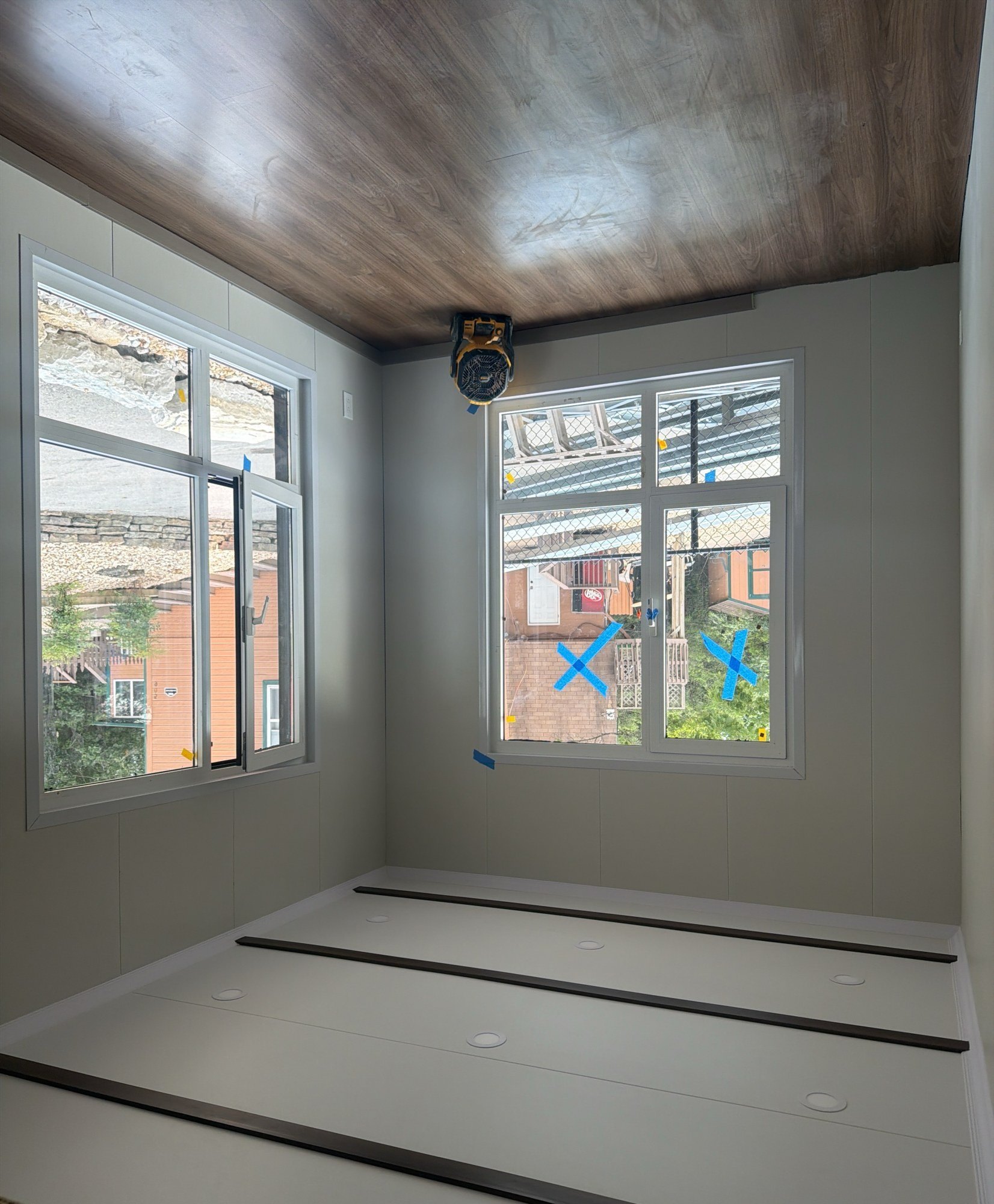 Finished bedroom with a wood-grain lined ceiling, white wall panels and two windows looking onto the site, 30 July 2026