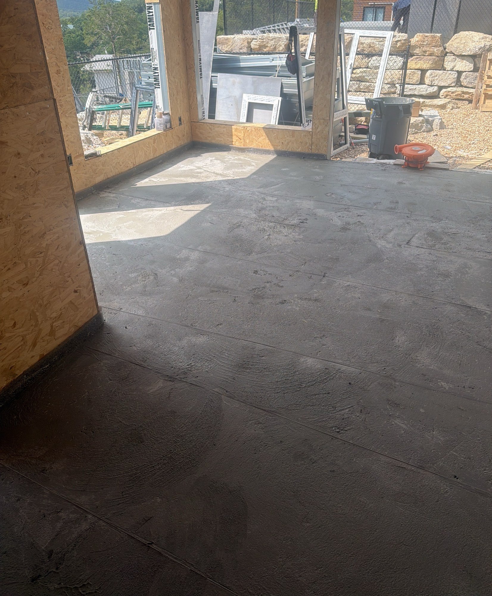 Finished grey floor topping in a sheathed room with window openings looking out to a stone retaining wall, 25 July 2026