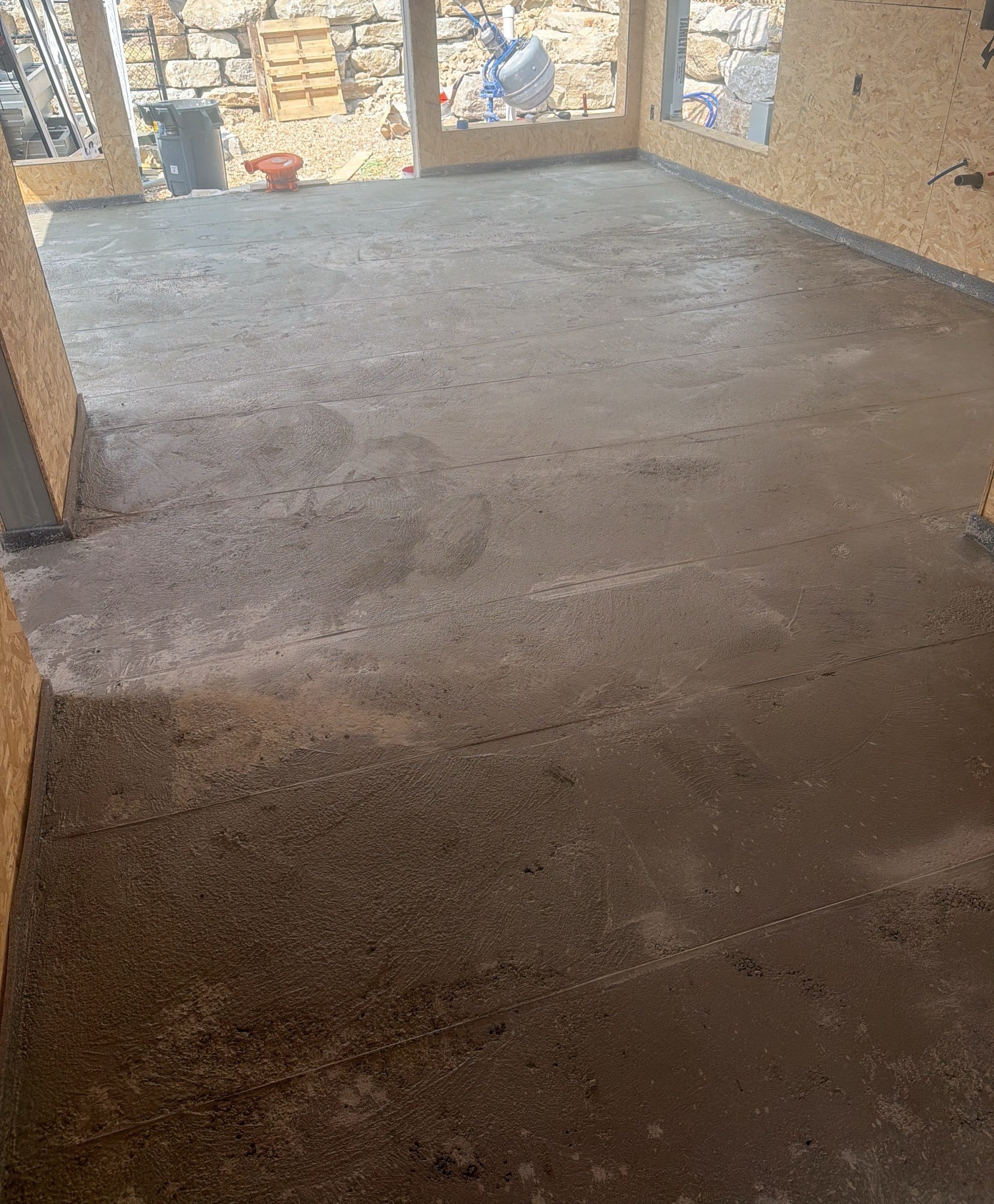 Levelled grey floor topping running across two sheathed rooms towards window openings, 25 July 2026