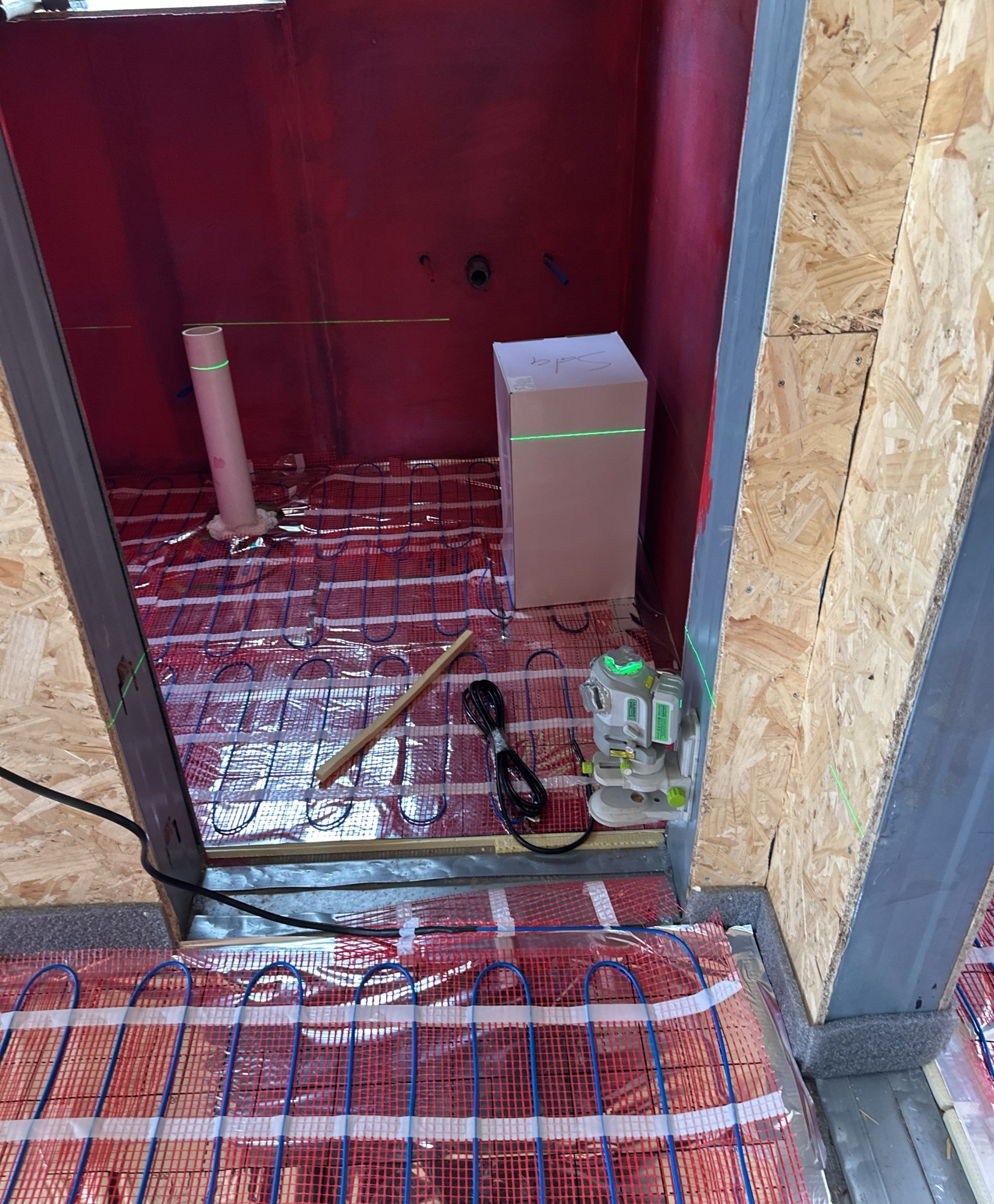 Electric underfloor heating cable laid in a serpentine pattern across a membrane mat in a bathroom, with pipe stubs standing at the far wall, 24 July 2026