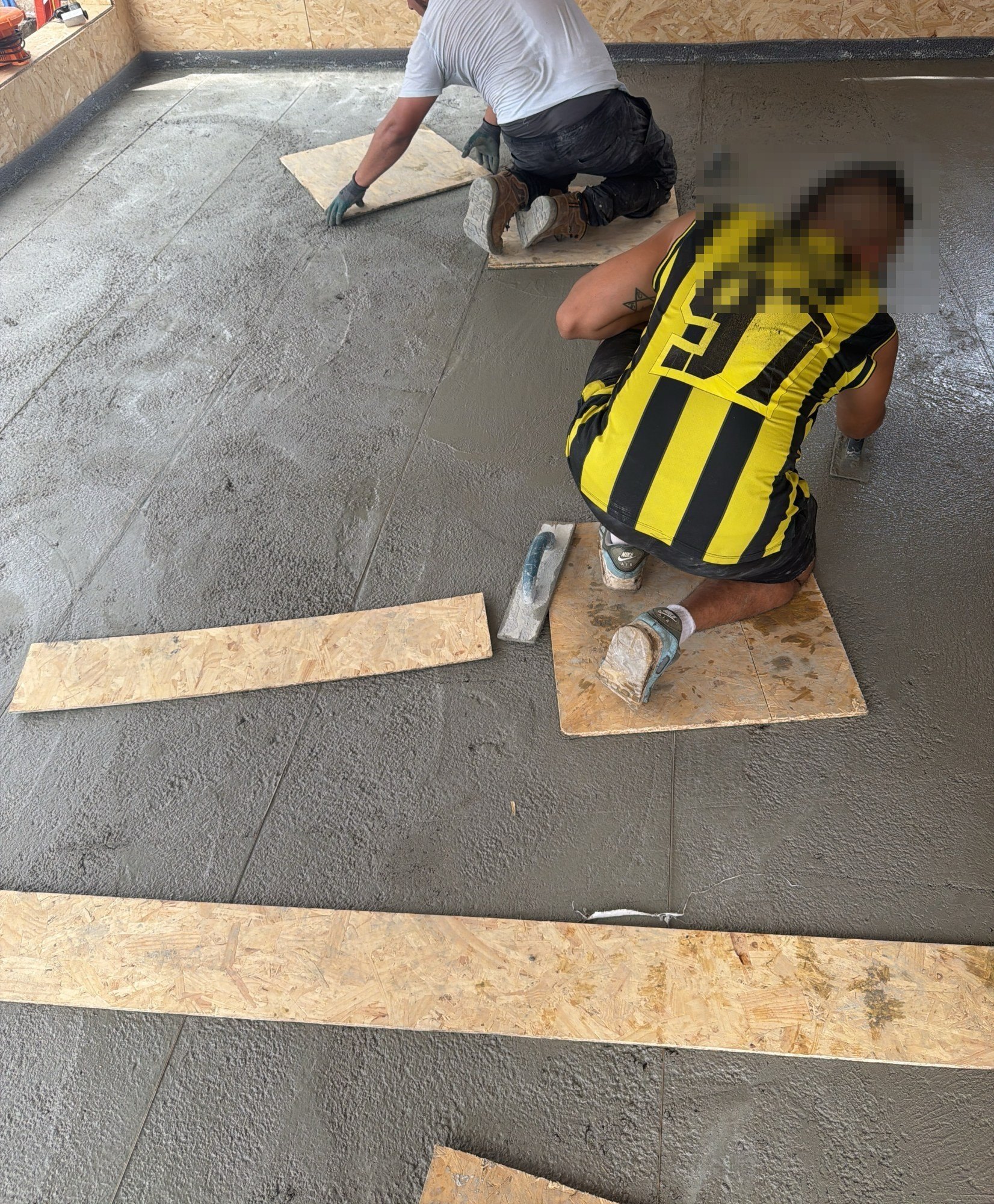 Worker kneeling on board offcuts while floating a grey topping compound across a floor, a second worker working behind, 25 July 2026