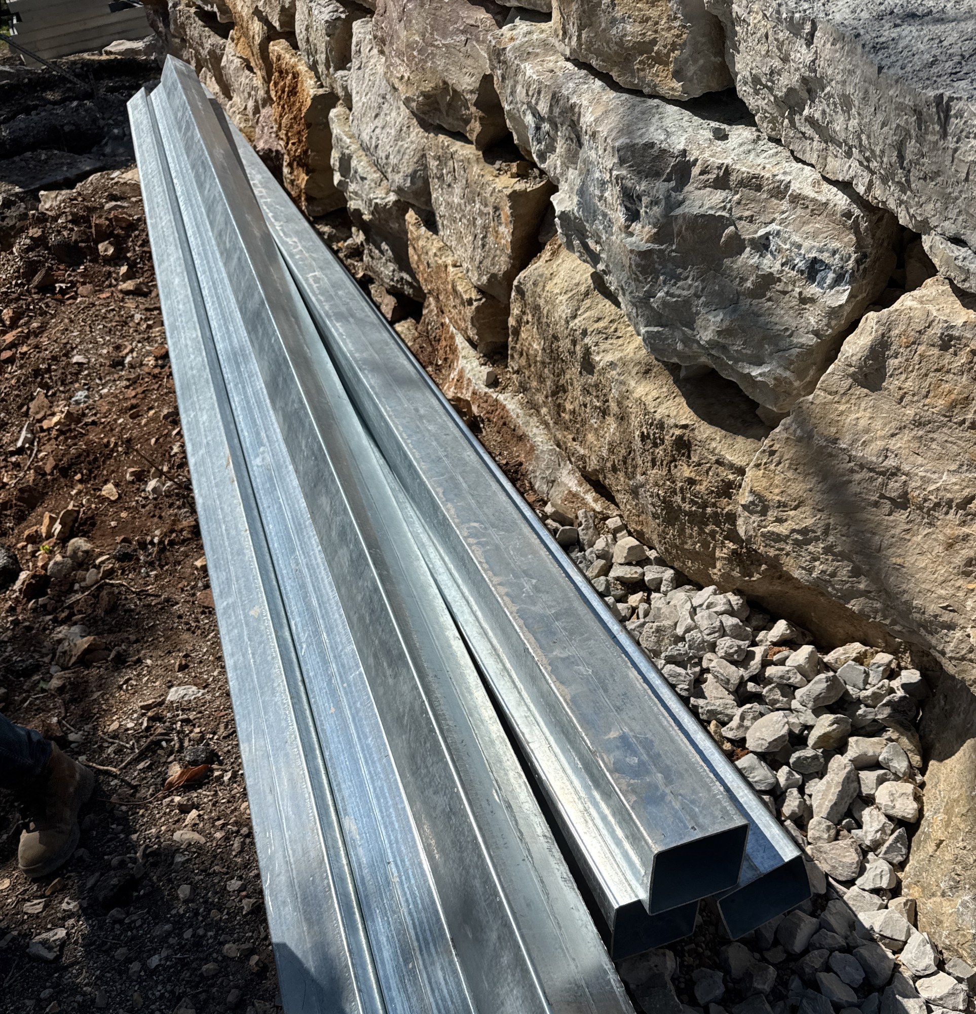 Galvanized cold-formed steel floor sections stacked on gravel beside a stone retaining wall, 4 May 2026